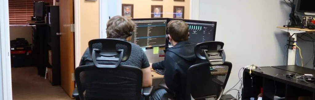 Two interns in headsets sit at a desk of dual monitors in the InfoWest tech-support room, their backs to the camera.