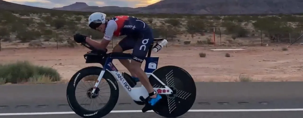 A triathlete on a time-trial bike rides a desert highway at dawn, red hills in the distance.