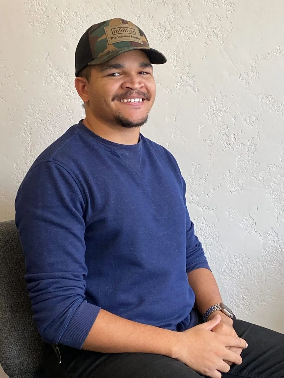 Gabriel Price sits smiling in a navy sweater and a camo InfoWest cap against a textured wall.