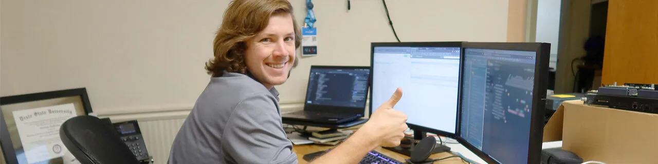 Jonathan sits at a desk of dual monitors and a laptop, turning to the camera with a thumbs up and a smile.