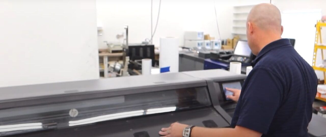 A man in a print shop operates a large-format HP printer, rolls of media and equipment behind him.