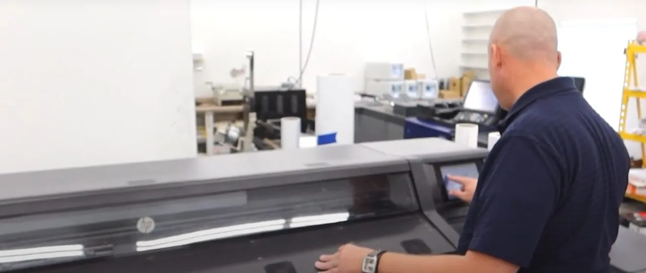 A man in a print shop operates a large-format HP printer, rolls of media and equipment behind him.