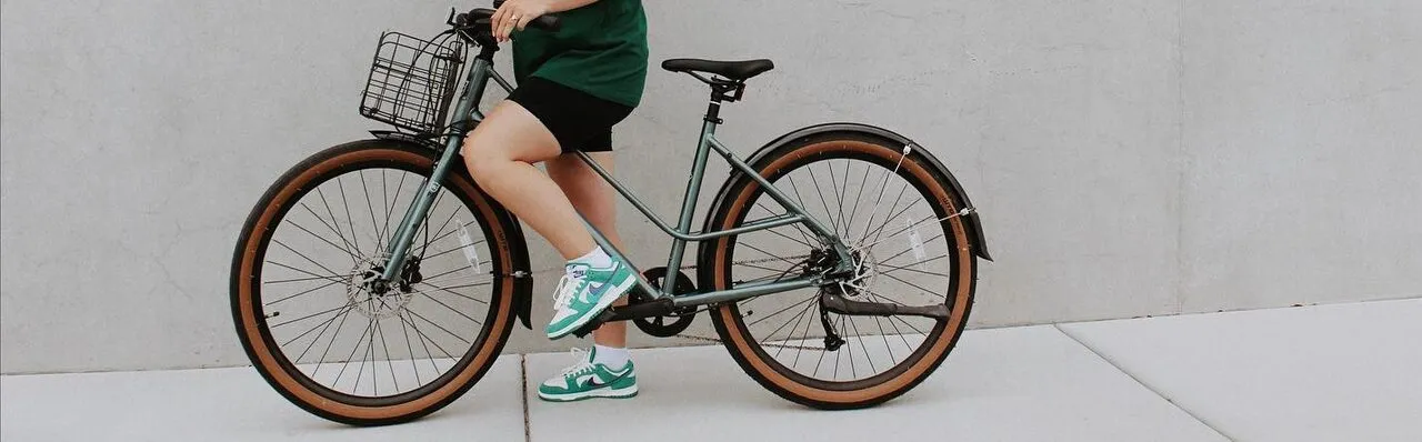 A person in shorts and sneakers sits on a teal cruiser bike with a front basket, against a plain wall.