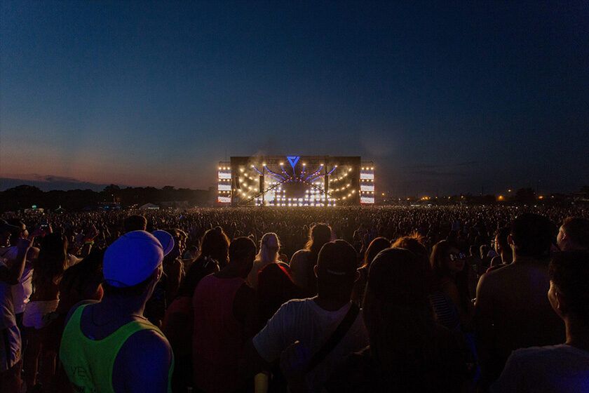 A large crowd at dusk facing a brightly lit outdoor concert stage