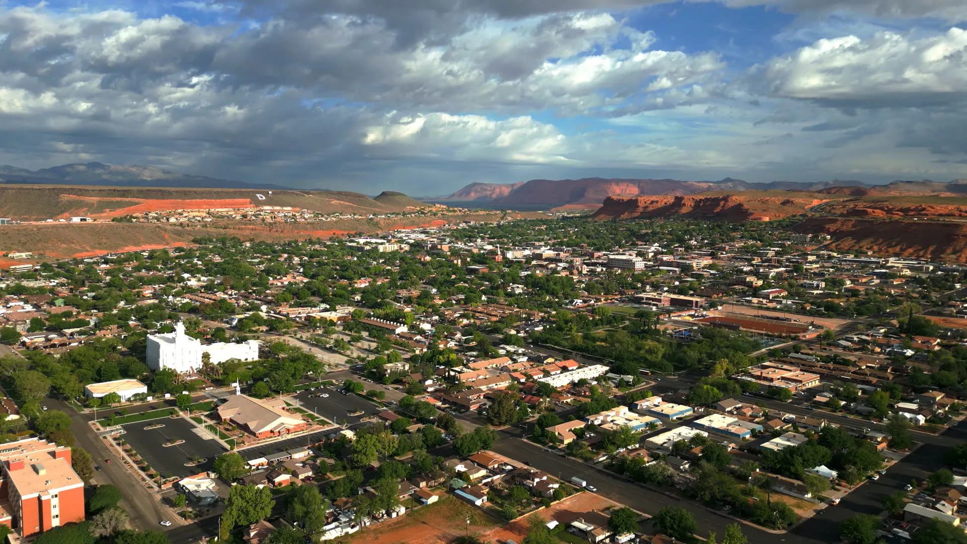 Aerial view of St. George at golden hour, neighborhoods and the temple spread below red mesas and a cloud-streaked sky.