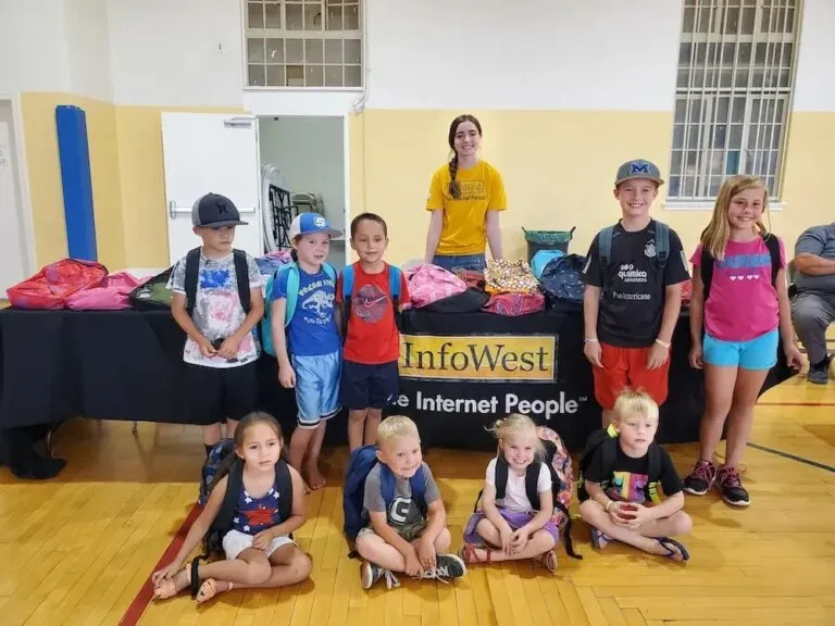 Children wearing new backpacks stand and sit around an InfoWest table in a gym at the backpack giveaway.
