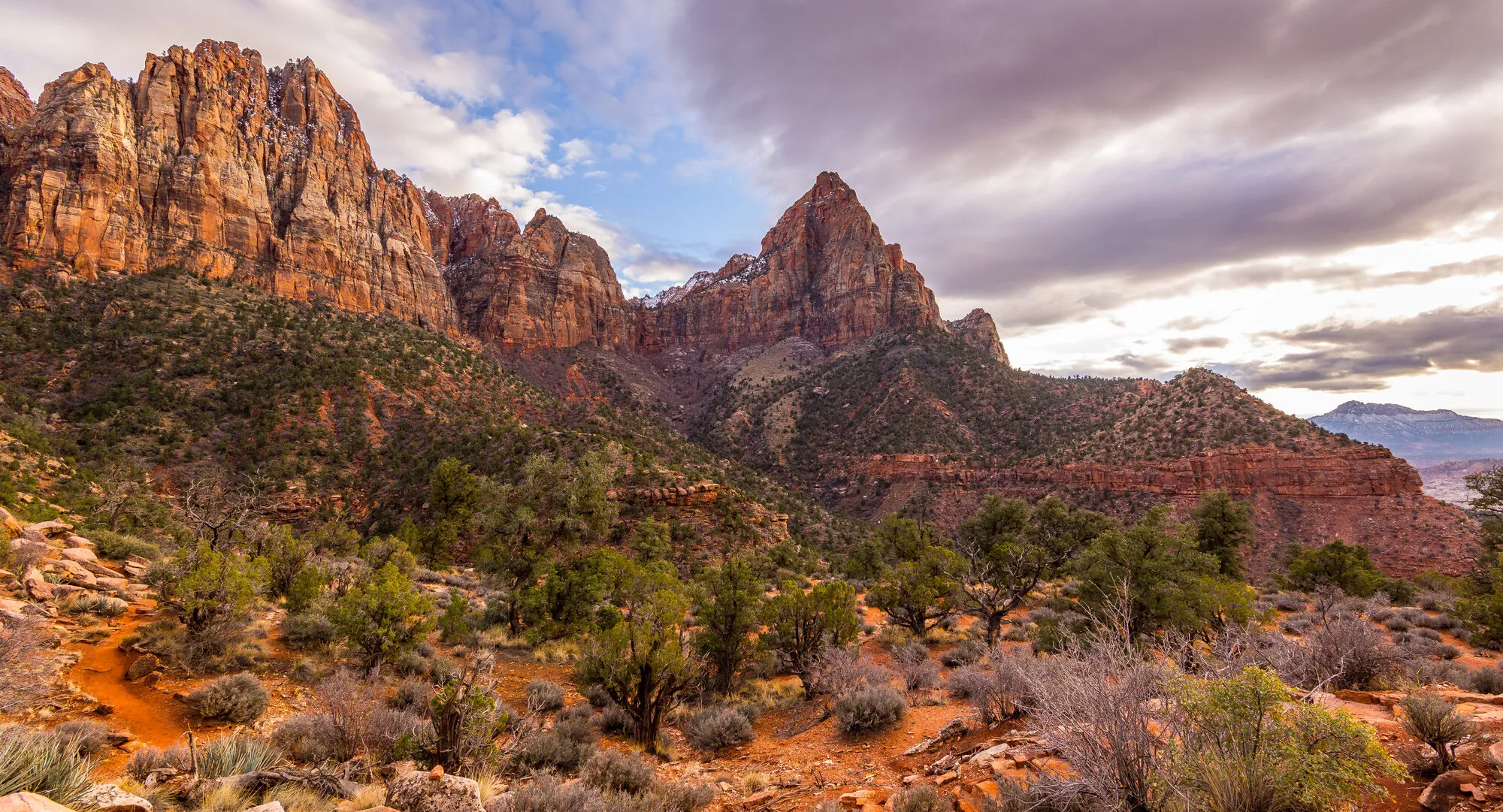 Red sandstone peaks and green juniper slopes near Zion National Park under a cloudy sky.