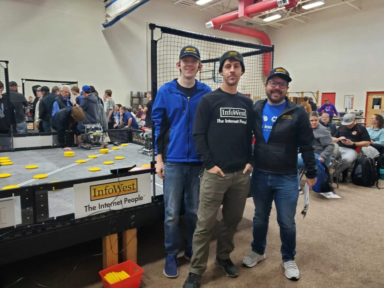 Three InfoWest volunteers beside a VEX Robotics competition field with an InfoWest sponsor sign